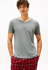 Camiseta gris de cuello en v en una tela suave, con mangas cortas y un pequeño logo. Combinada con pantalones de pijama a cuadros rojos, que muestran un patrón de cuadros.