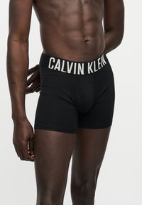 Αρσενικός κορμός που φοράει μαύρα μποξεράκια Calvin Klein με ορατό λογότυπο στην ελαστική ζώνη, σε απλό λευκό φόντο.