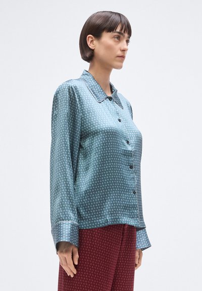 Niebieska jedwabna koszula z kołnierzykiem, ozdobiona drobnym białym wzorem i czarnym wykończeniem na mankietach i kołnierzu. Zapięcie na guziki z przodu.