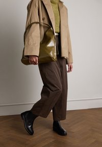 Giacca leggera beige, borsa in pelle verde oliva, maglione strutturato senape, pantaloni a gamba larga marroni, stivali neri alla caviglia con suola spessa.
