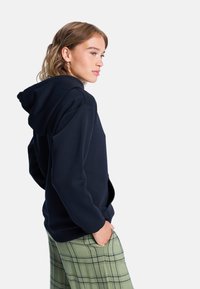 Sweat-shirt à capuche bleu marine en tissu doux. Présente une coupe décontractée et une poche kangourou. Associé à un pantalon à carreaux vert clair.