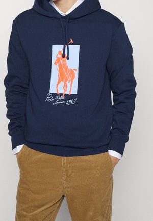 Mężczyzna w granatowej bluzie z kapturem z pomarańczową grafiką gracza polo i napisem "Polo Ralph Lauren 1967", w połączeniu z jasnobrązowymi sztruksowymi spodniami.