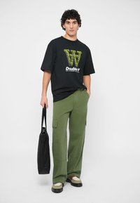 Črna majica s grafičnim zelenim napisem, olivno zelene cargo hlače, bež in črni čevlji ter črna torba preko rame, stoji pred belo ozadjem.