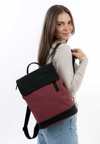Sac à dos bordeaux et noir avec un design à rabat, fabriqué en matériau durable, doté d'une sangle ajustable et d'une poignée supérieure pour le transport.