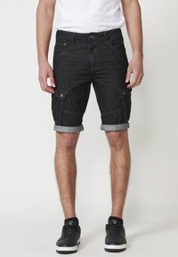 Schwarze Denim-Cargo-Shorts mit umgeschlagenen Säumen, ausgestattet mit zwei knöpfbaren Seitentaschen und Stickdetails, getragen mit schwarzen Sneakers.