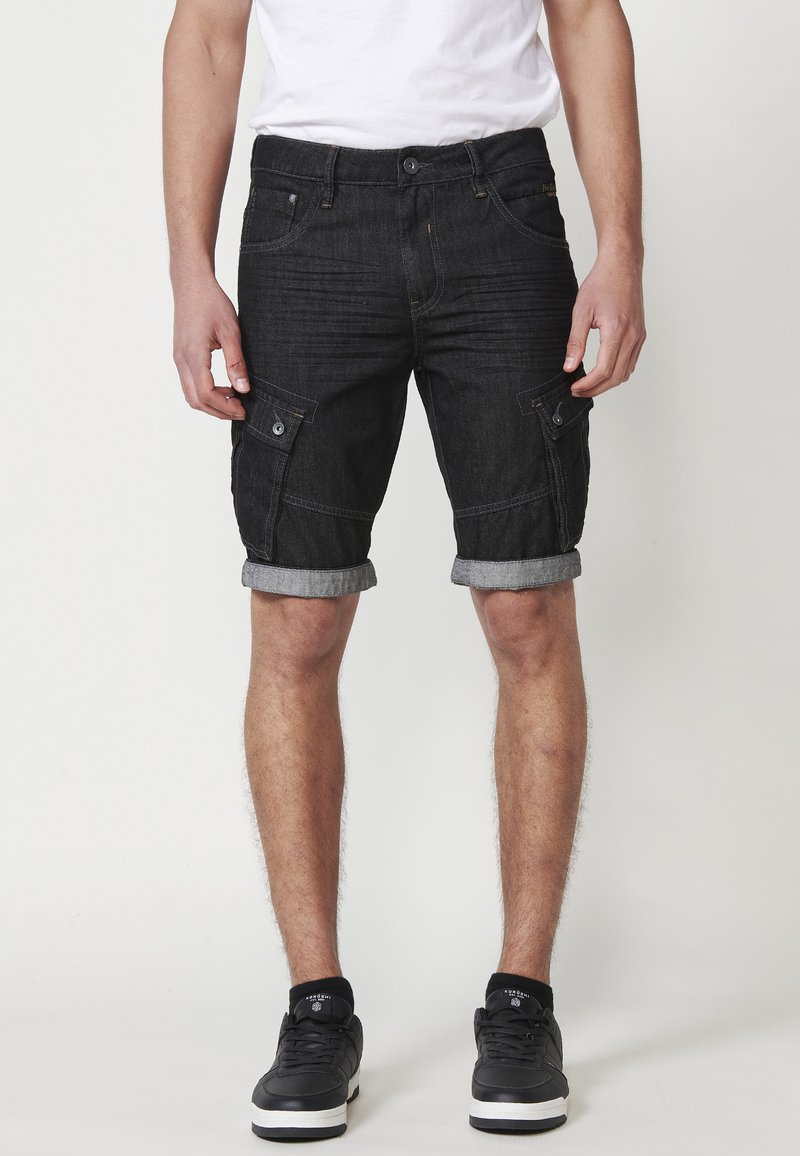 Schwarze Denim-Cargo-Shorts mit umgeschlagenen Säumen, ausgestattet mit zwei knöpfbaren Seitentaschen und Stickdetails, getragen mit schwarzen Sneakers.
