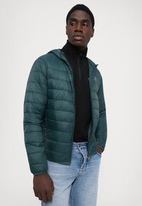 Mørkegrøn pufferjakke med hætte, prydet med vandrette quiltede linjer. Bæres over en sort lynlåsbluse og lyseblå jeans.