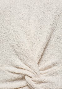 Gros plan sur un tissu doux de couleur blanc cassé, texturé et torsadé en nœud, mettant en valeur sa matière en boucle, semblable à une serviette, et son aspect confortable.