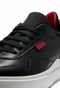 Mustat nahkakengät, joissa on teksturoitu pinta, tasaiset mustat nauhat, punainen logo-etiketti ja valkoinen kumipohja. Sisäosa on punainen.