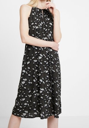 Vrouw draagt een mouwloze zwarte midi-jurk met grijze en witte abstracte vlekken, staat met een hand dicht bij haar gezicht en de andere over haar taille.