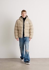 Young man walking indoors wearing a beige puffer jacket, black shirt, ripped blue jeans, and black-and-white sneakers.