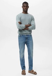 Lichtblauwe trui met ronde hals, blauwe jeans en bruine sandalen. De trui heeft een geribbelde halslijn en een ontspannen pasvorm; de jeans hebben een klassieke snit.