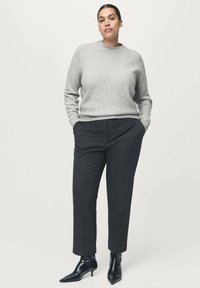 Frau mit zurückgehaltener dunkler Haare, die einen hellgrauen Pullover, schwarze cropped Hose und schwarze spitze Absatzstiefel trägt, steht mit den Händen in den Taschen.