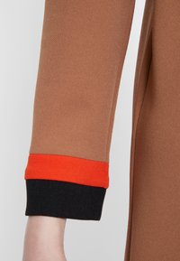 Braunes Langarmkleidungsstück aus strukturiertem Stoff, mit einem orange und schwarzen gestreiften Bündchen am Handgelenk.