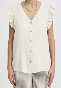 Frau trägt eine ärmellose, cremefarbene Bluse mit Knöpfen und gerafften Schultern sowie gestreifte beige und schwarze Hose, ergänzt durch eine goldene Halskette.