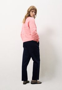 Rosa strukturierter Pullover mit lockerer Passform, kombiniert mit marineblauen Hosen. Die Schuhe haben ein Leopardenmuster. Der Hintergrund ist schlicht und neutral.