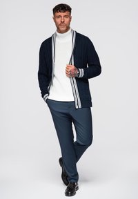 Cardigan navy con lavorazione a treccia e dettagli a strisce bianche, indossato su un dolcevita bianco, abbinato a pantaloni blu scuro e mocassini neri.