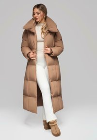 Žena v dlouhém hnědém péřovém kabátu, béžovém roláku, bílých kalhotách a semišových boots s kožíškem, stojící na jednolitém šedém pozadí.