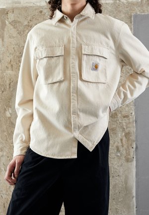 Osoba nosząca kremową koszulę zapinaną na guziki z dwoma kieszeniami na piersi i naszywką z logo Carhartt, w połączeniu z czarnymi spodniami na tle teksturowanej ściany.