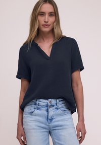 Femme aux longs cheveux blonds portant un chemisier texturé bleu marine foncé à manches courtes et un jean taille haute bleu clair, debout devant un fond uni.
