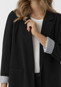 Woman wearing a black blazer with striped cuffs over a white ribbed shirt, holding the lapel with her right hand.