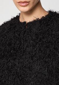 Giacca nera con texture che presenta una finitura pelosa, scollatura rotonda e chiusura frontale con cerniera. Il materiale appare morbido con lunghezze di fibra variabili.