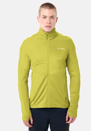 Jonge man met een limoengroene jas met ritssluiting en duimgaten, staande tegen een effen lichte achtergrond, recht vooruit kijkend.