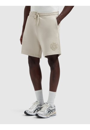 Mann trägt beige Shorts mit Kordelzug und einem aufgestickten Logo am linken Bein, weiße Sportsocken und silberne Laufschuhe.