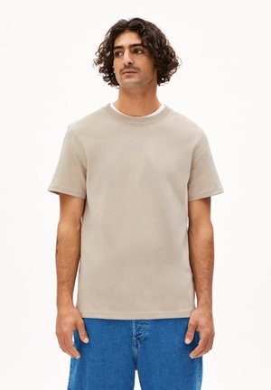 Beige Baumwoll-T-Shirt mit Rundhalsausschnitt und kurzen Ärmeln, das eine glatte Textur und einen lässigen Schnitt aufweist. Getragen mit blauen Denim-Unterteilen.