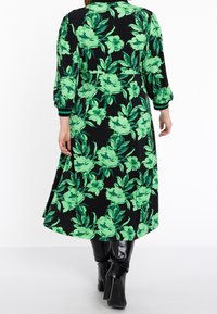 Musta mekko, jossa on iso vihreä kukkakuosi, pitkät hihat ja joustavat hihansuut. Mekossa on virtaava siluetti ja polviin ulottuva helma.