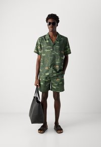 Grøn kortærmet skjorte og shorts sæt med et tropisk print. Sorte solbriller og en stor sort toilettaske. Flade sandaler fuldender outfittet.