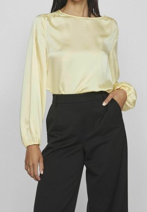 Femme portant un blouse en satin jaune pâle à manches longues avec des poignets élastiques, rentrée dans un pantalon noir taille haute, main dans la poche.