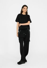 Schwarzes T-Shirt mit kurzen Ärmeln und strukturiertem Detail, kombiniert mit schwarzen, weit geschnittenen Hosen mit seitlichen Taschen und weißen Paspeln.