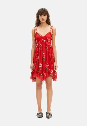 Donna che indossa un vestito senza maniche rosso a fiori con volant e spalline annodate, abbinato a ballerine nere con borchie, in piedi davanti a uno sfondo uniforme.