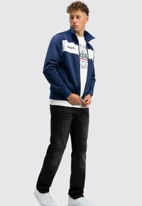 Junger Mann in einer marineblauen und weißen Reißverschlussjacke, einem weißen T-Shirt mit Grafik, schwarzen Jeans und weißen Turnschuhen, der vor einem hellen Hintergrund steht.