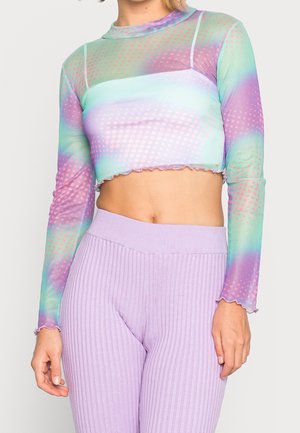Vrouw draagt een langemouwige, doorschijnende crop top met pastelgroene, paarse en roze patronen, gecombineerd met hooggetailleerde geribbelde lavendelkleurige broek.