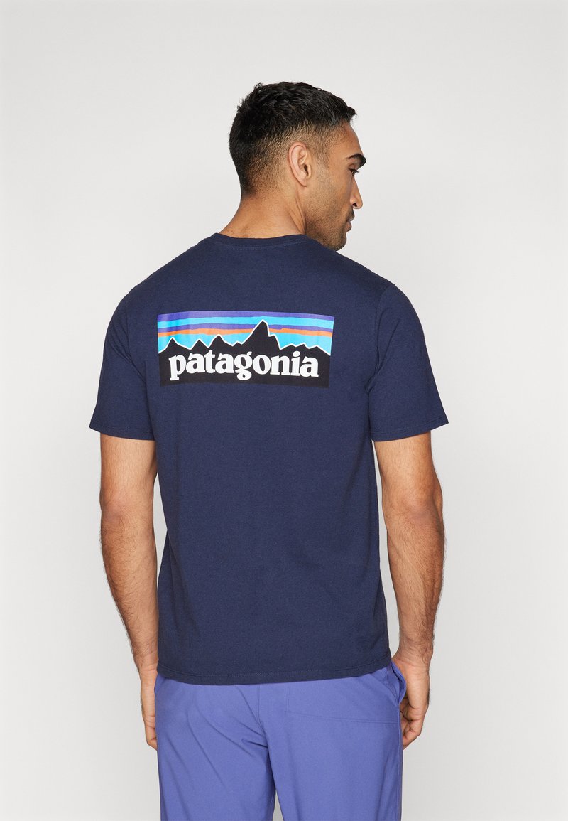 Tricou din bumbac de culoare albastru închis, cu un logo colorat Patagonia și un grafic cu munți pe spate. Mâneci scurte și decolteu rotund.