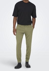 T-shirt noir à manches courtes associée à un pantalon slim vert olive. Le pantalon présente une texture lisse et des poches avant. Des bottines noires complètent la tenue.