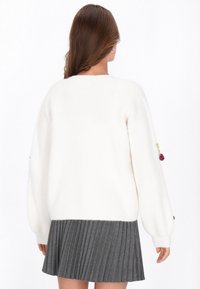 Witte gebreide trui met ribbeltextuur en oversized mouwen. Het heeft kleurrijke geborduurde accenten op de mouwen. Gecombineerd met een geplooide grijze rok.
