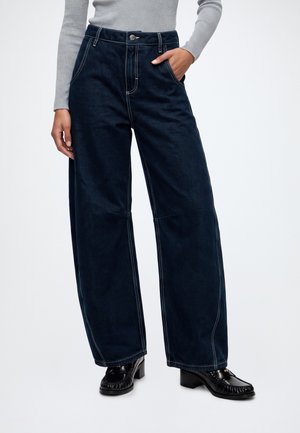 Personne portant un jean large foncé, un haut gris côtelé à manches longues et des mocassins noirs à talons, debout avec une main dans la poche.