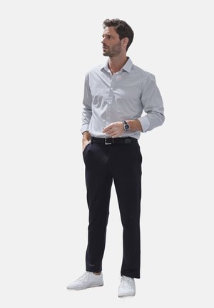 Camicetta a righe bianche e blu navy a maniche lunghe, pantaloni neri, cintura argentata e sneakers bianche. Indossa un orologio da polso; in piedi con le mani nelle tasche.