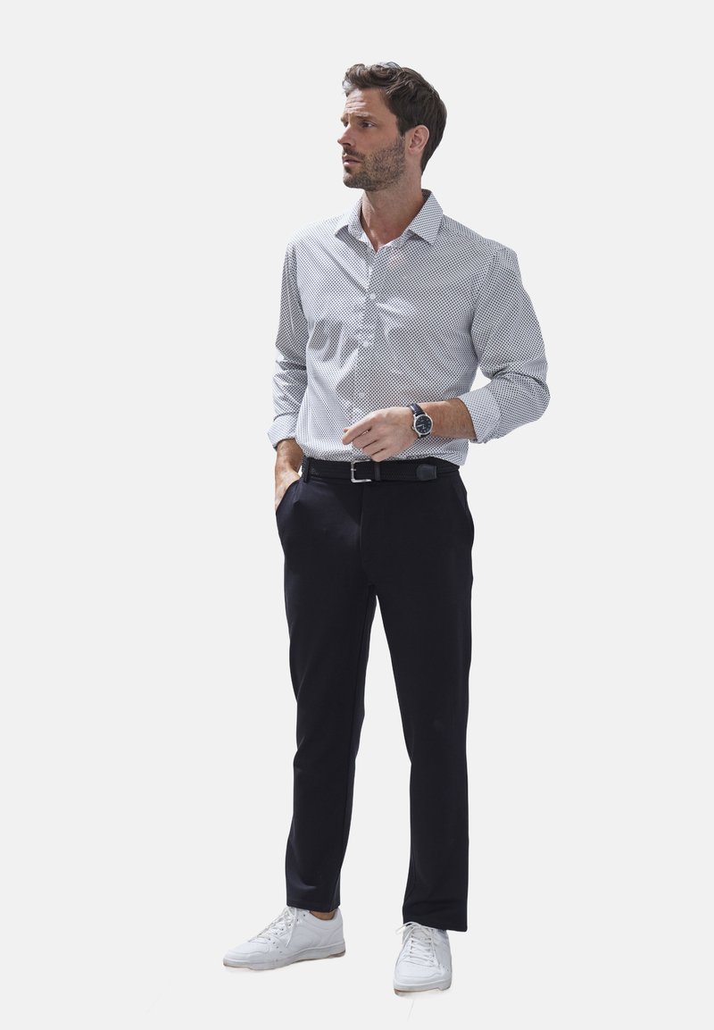 Wit en marineblauwe gestreepte lange mouwen shirt, zwarte broek, zilveren riem en witte sneakers. Draagt een polshorloge; staat met de handen in de zakken.