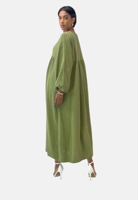 Olivegrünes Maxikleid aus strukturiertem Stoff, mit langen Puffärmeln und Empire-Taille. Kombiniert mit weißen High-Heels.