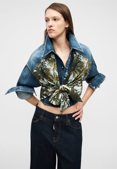 Γυναίκα που φορά πουκάμισο από τζιν με τυπωμένο τροπικό μοτίβο με τα μανίκια γυρισμένα και δεμένο στη μέση, καθώς και σκούρο τζιν, ποζάροντας με τα χέρια στη μέση.