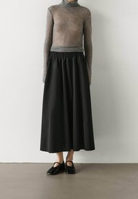 Maglione grigio a collo alto abbinato a una gonna nera lunga fino alla caviglia con vita elasticizzata e scarpe nere basse dal design minimal.