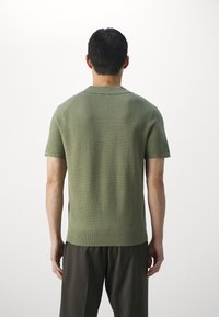 Vihreä neulottu lyhythihainen neule teksturoidulla kuviolla, resorikauluksella ja sivuhalkioilla, tummien housujen kanssa, nähtynä takaa.