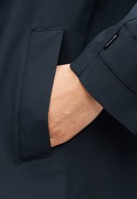 Χέρι που έχει εισαχθεί στην τσέπη σκούρου μπλε παλτού με ορατή ετικέτα υφάσματος «Ανακυκλωμένο» στο μανίκι.