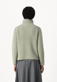 Pulover ribbed de culoare verde deschis, cu guler înalt, siluetă ajustată și mâneci lungi. Purtați peste o fustă gri, textura este moale și texturată.