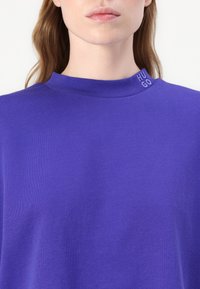 Top corto viola a maniche corte con scollo alto. Presenta il marchio "HUGO" al colletto, realizzato in un tessuto liscio e leggero.