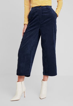 Personne portant un pantalon large court en velours côtelé bleu marine, un haut à manches longues rayé jaune et blanc, et des bottines blanches à talons.